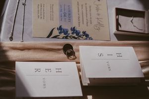 Wedding flat lay with floral invitation, vow booklets reading HE and SHE YOURS, gold rings, and elegant stationery on silk fabric.