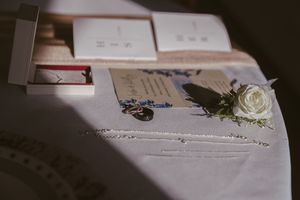 Wedding flat lay with white rose boutonniere, invitation cards, pearl necklace, and ring box on white linen table.