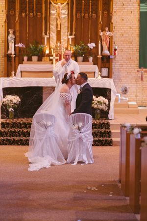 Bride and groom share first kiss at church altar during Catholic wedding ceremony with priest officiating.