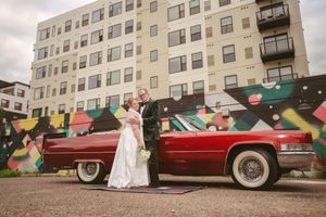 Bride and groom posing beside a classic red Cadillac in front of a colorful mural and modern building.