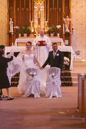 Newlyweds hold hands at Catholic church altar with priest officiating wedding ceremony behind decorated white chairs.