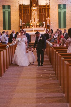 Bride and groom walk down church aisle with flower girl after wedding ceremony, guests watching from wooden pews.