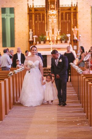 Bride and groom walk down church aisle with young flower girl between them during wedding ceremony.