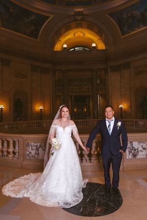 A bride in a lace gown and groom in navy suit hold hands inside an ornate domed wedding venue.