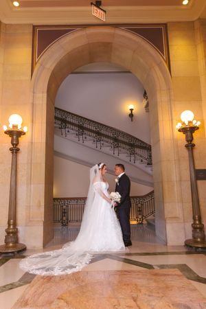 Bride and groom share a romantic moment under an elegant arch with grand staircase backdrop.