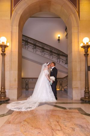 Bride and groom share romantic kiss under elegant archway with grand staircase and ornate lamps.