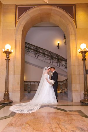 Bride and groom share romantic moment under grand arch with ornate staircase and golden candelabras in elegant venue.