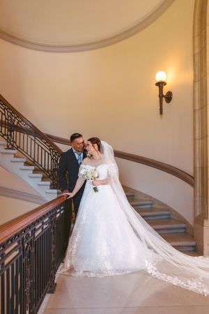 Wedding couple embraces on curved staircase with iron railing, bride in flowing lace gown with long veil.