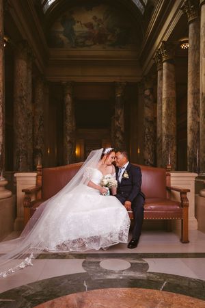 Bride and groom share a romantic moment on a bench in an elegant marble hall with ornate columns and painted ceiling.