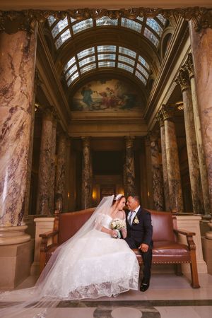 Bride and groom share intimate moment on ornate bench in grand hall with marble columns and painted ceiling.