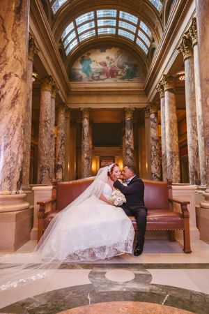 Bride and groom share a romantic moment on a bench inside an ornate marble hall with tall columns and painted ceiling.