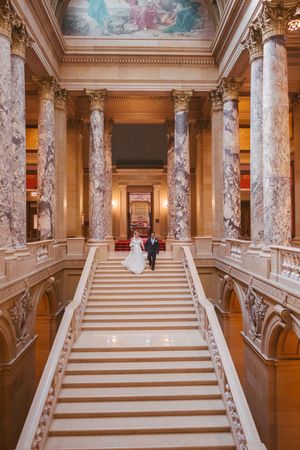 A bride and groom walk down a grand marble staircase beneath a classical painted ceiling mural.