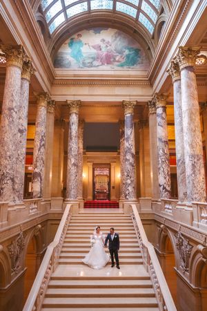 Wedding couple poses on grand marble staircase beneath stunning ceiling mural in ornate historic building.