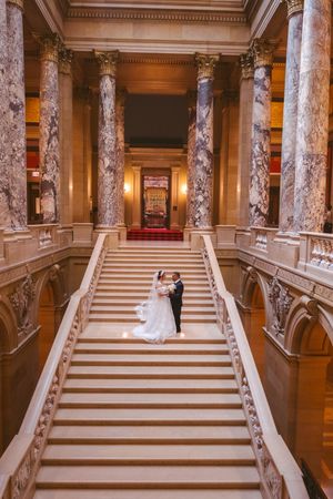 Bride and groom stand on grand marble staircase flanked by towering columns in stunning neoclassical building.