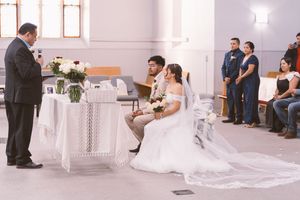 A bride and groom sit together during a church wedding ceremony while an officiant reads vows at the altar.