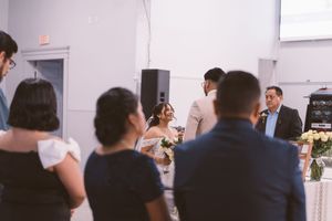 Wedding reception guests watching bride and groom share a moment near a floral arrangement in a bright venue.