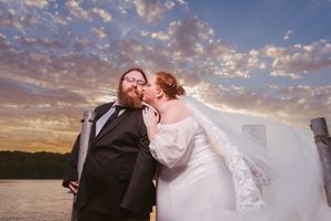 Bride and groom share a romantic kiss at sunset by the water, with dramatic clouds filling the sky behind them.