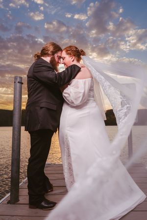 Bride and groom share romantic sunset moment on dock, veil flowing in wind, with dramatic cloudy sky over water.