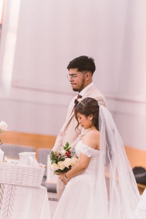 A bride and groom stand at the altar during their wedding ceremony, she holds a bouquet of white and red roses.