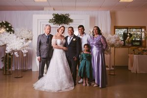 Wedding party poses in decorated venue with bride in white gown, groom, family members, and girl in green dress.