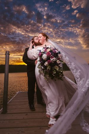 Bride and groom share a romantic kiss on a dock at sunset, with a flowing veil and colorful bouquet.