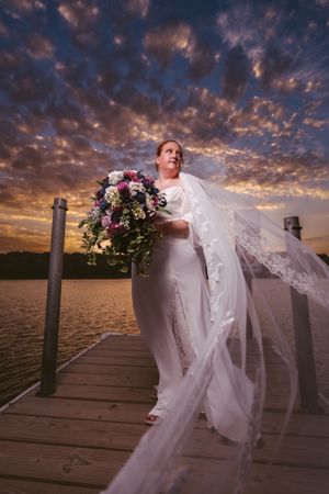 Bride in white gown holding floral bouquet on dock at sunset with dramatic cloudy sky.