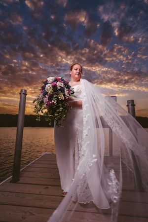 Elegant bride with flowing veil holding bouquet on waterfront dock beneath vibrant sunset sky.