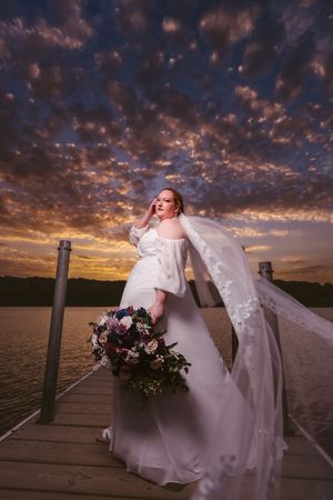Bride in white off-shoulder gown holding bouquet on dock at sunset with dramatic cloudy sky over water.