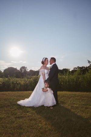 Bride and groom share a romantic moment outdoors at golden hour, with a small child between them on lush green grass.