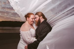 Bride and groom share a romantic moment on a beach at sunset, with the wedding veil billowing around them.