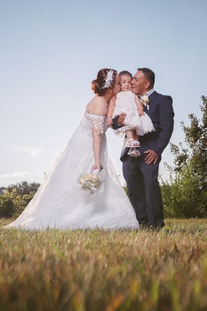 Bride and groom in wedding attire kiss their young daughter outdoors on a sunny day in a lush green field.