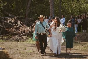 Bride in white gown walks with man in cowboy hat and child in green dress at outdoor woodland wedding ceremony.