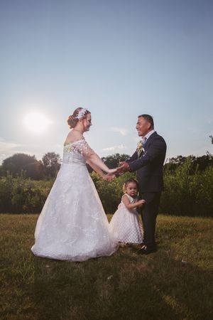 Happy couple in wedding attire holding hands outdoors with a young flower girl between them at sunset.