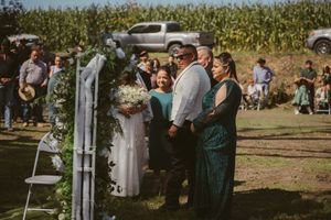 Outdoor wedding ceremony at a farm with couple and guests standing under a floral arch near a cornfield.