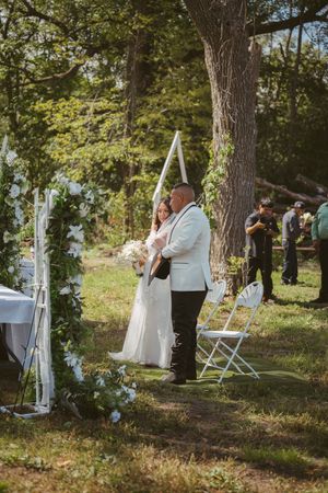 Bride and groom embrace during outdoor wedding ceremony surrounded by white floral arch and lush green trees.
