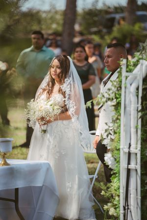 Bride in lace wedding dress and veil holds white bouquet at outdoor ceremony with guests and greenery.