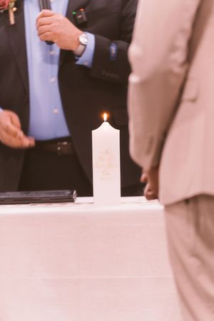 A lit white unity candle on a draped table during a wedding ceremony with guests standing nearby.