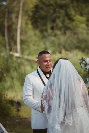 Groom in white tuxedo smiling at bride in lace veil during outdoor wedding ceremony.