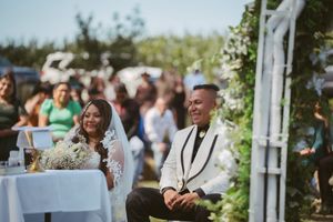 Smiling bride and groom seated outdoors at a sunny wedding ceremony with floral arch and guests in background.
