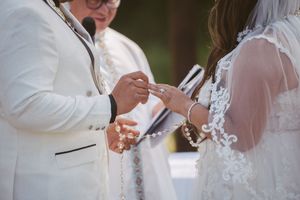 Groom in white suit placing wedding ring on bride's finger during outdoor ceremony with officiant.