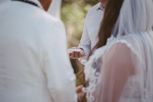 Close-up of a wedding ceremony ring exchange, bride in lace veil and groom in gray shirt outdoors.