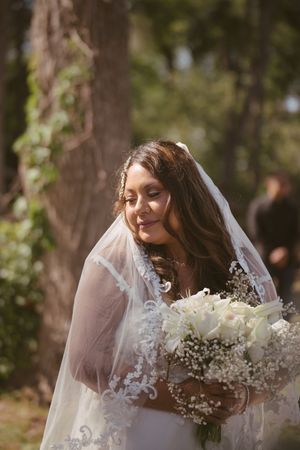 Smiling bride in lace wedding dress and veil holding white floral bouquet outdoors among trees.