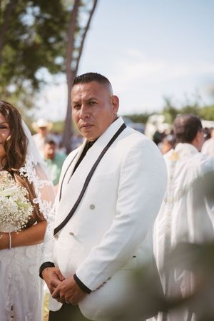 Groom in white black-trimmed tuxedo stands attentively at outdoor wedding ceremony.