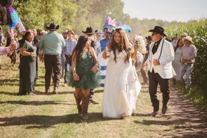 Bride walks with wedding party in boho outdoor ceremony, guests in cowboy hats line rustic path.