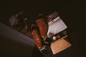 Brown leather oxford shoes with laces beside a watch, wallet, and accessories arranged on a dark surface.