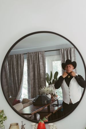 Man in cowboy hat and vest adjusting collar, reflected in round mirror with home interior background.