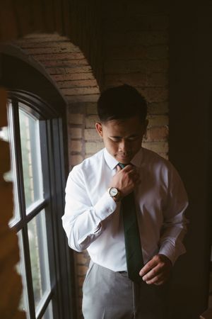 Groom in white shirt adjusting green tie by window with brick wall backdrop on wedding day.