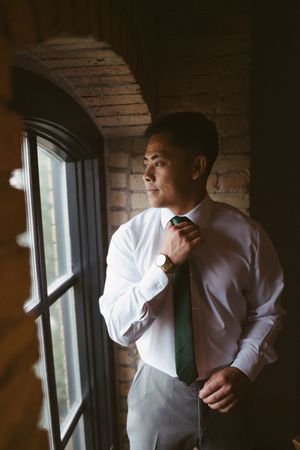 Groom in white shirt adjusting green tie by arched window in brick wall venue before wedding ceremony.