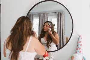Bride in white dress adjusting hair accessories while looking in round mirror.