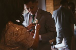 A woman pins a boutonniere on a groom's lapel during wedding preparations in warm indoor lighting.
